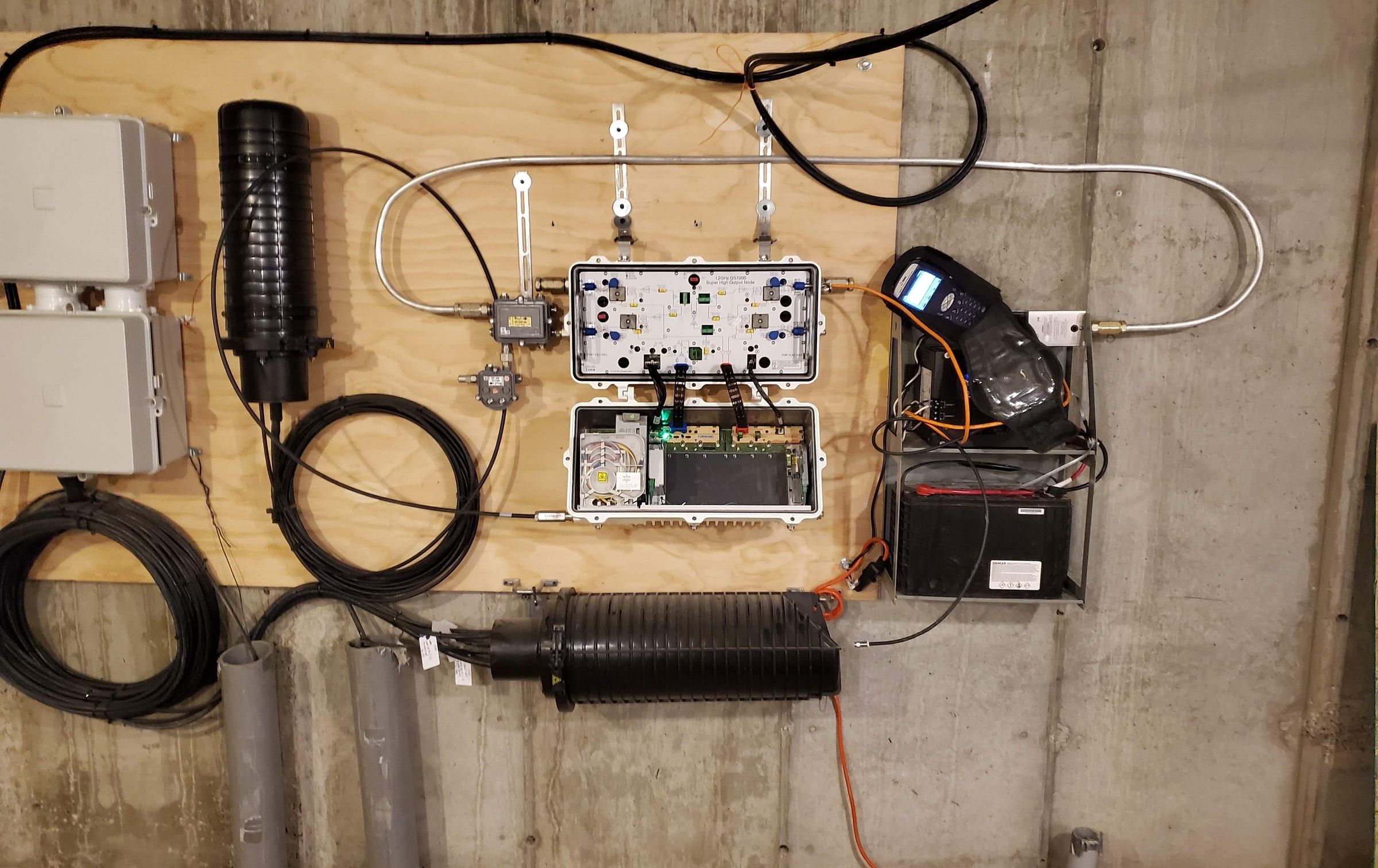 Wall-mounted node with splice closures, coiled cable and a signal meter on a plywood backboard
