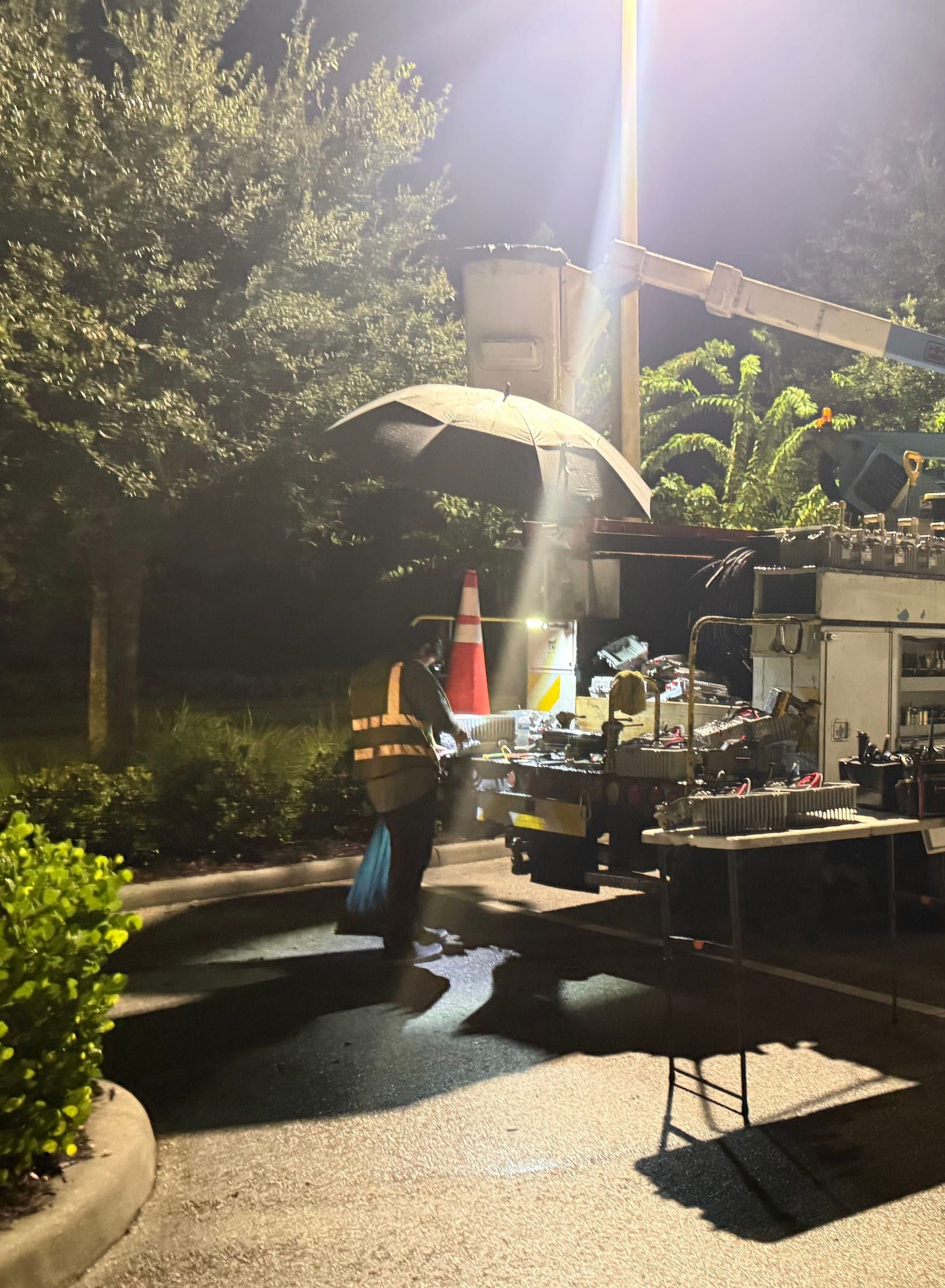 Night work under a bucket truck light, a crew member in a safety vest at a folding table of equipment